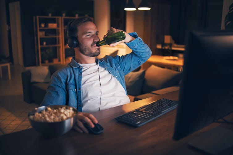 Young man drinks a bottle of beer with a snack at night in front of his computer.