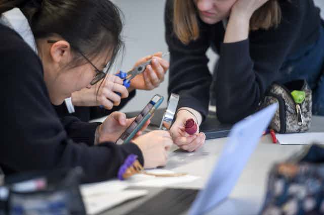 Three schoolgirls in uniform use a smartphone in an art class