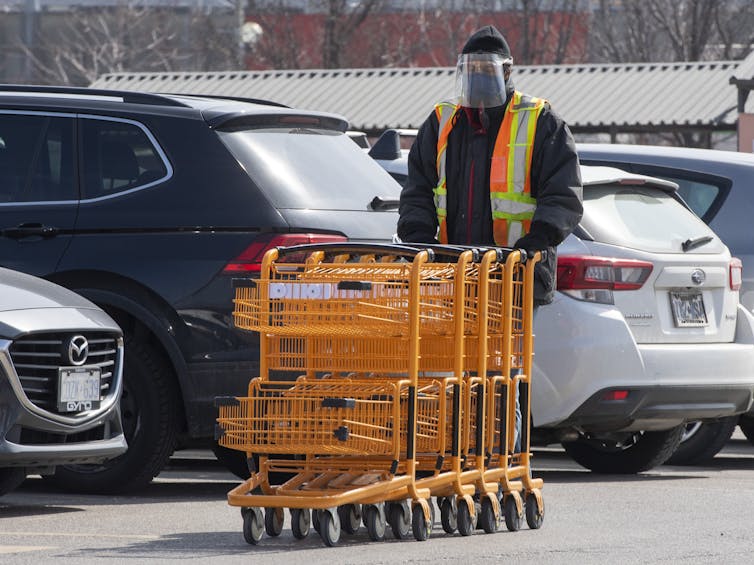 A person in a face shield pushing a row of shopping carts in a parking lot