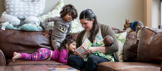 A woman reads to two young children while cradling a baby in her arm while their dog clambers on the couch.