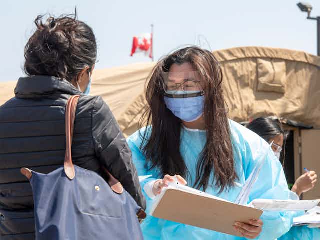 A health-care worker in personal protective equipment talks to a woman wearing a mask at an outdoor vaccine clinic.