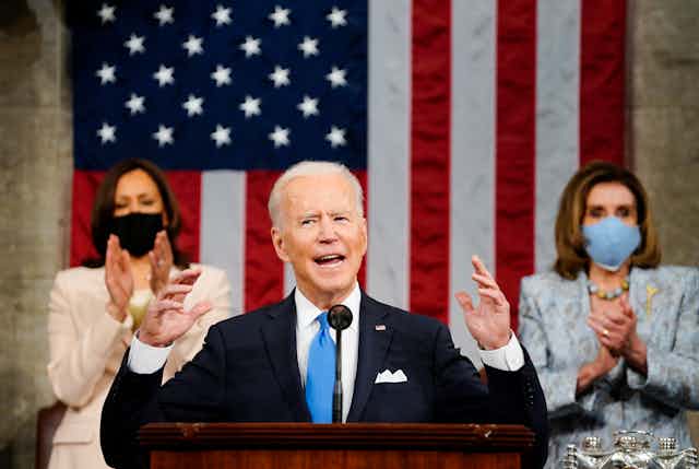 President Biden gesticulates with both hands as he speaks before Congress, flanked by Vice President Kamala Harris to his right and House Speaker Nancy Pelosi to his lift and an American flag draped behind him.