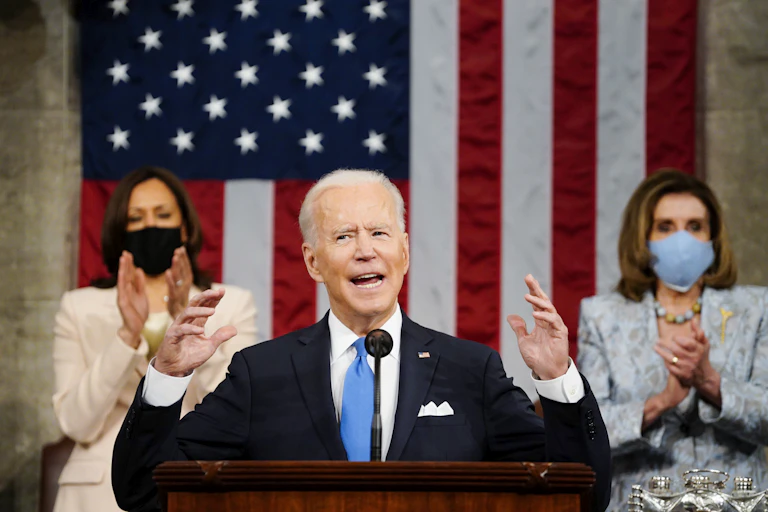 President Biden gesticulates with both hands as he speaks before Congress, flanked by Vice President Kamala Harris to his right and House Speaker Nancy Pelosi to his lift and an American flag draped behind him.