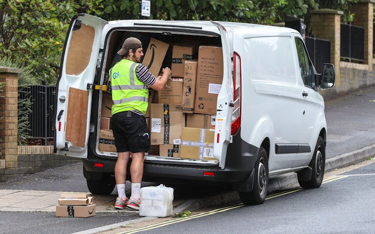 A delivery driver offloads boxes from a white van parked on the pavement