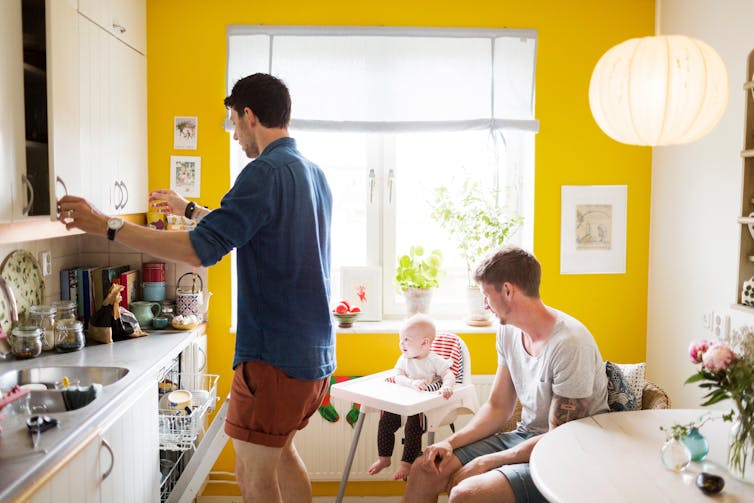 A gay couple prepare food in a yellow kitchen as a baby in a high chair looks on