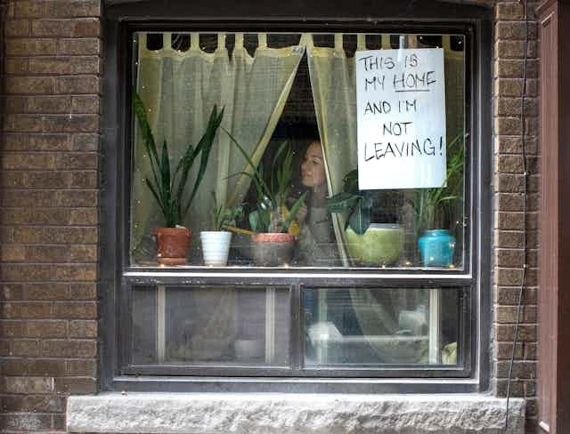 A woman looks through a window with a sign taped to it reading This Is My Home and I'm Not Leaving.