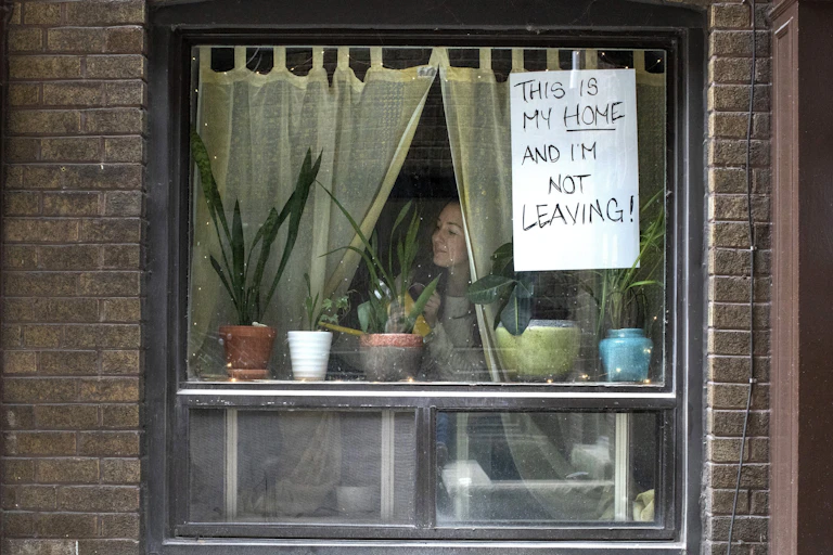 A woman looks through a window with a sign taped to it reading This Is My Home and I'm Not Leaving.