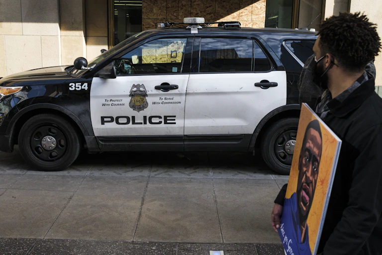 A man carrying a portrait of George Floyd looks on at a police cruiser as demonstrators march in honor of George Floyd.
