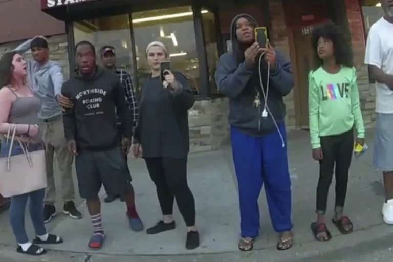 People stand on street corner watching the police. One woman is filming with her cell phone.