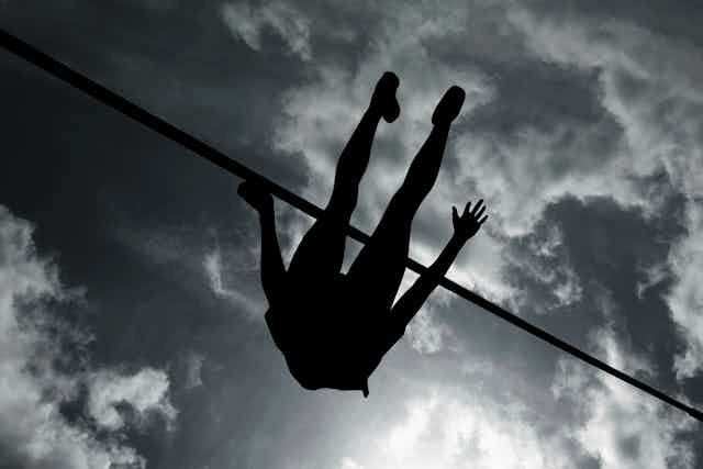 A silhouette of a body flying over a pole, shot from below, clouds in the sky above the high jumper.