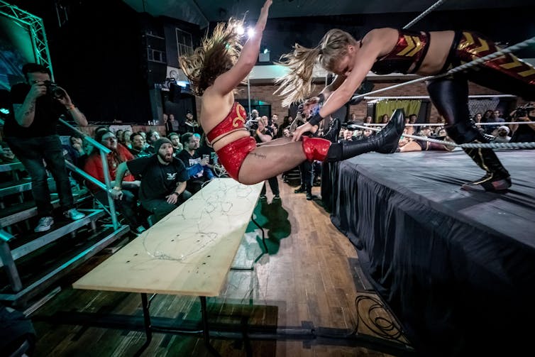 Female wrestler slams opponent into table from inside the wrestling ring
