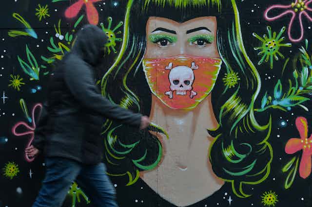 A man in a hoodie walks past a mural of a woman with green hair who wears a mask with a skull and crossbones over the mouth area.