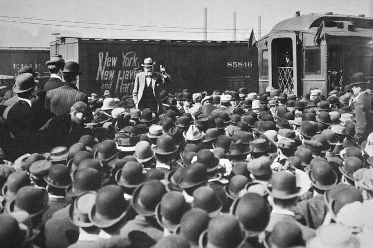 Black-and-white image of a man speaking from a train to a crowd of men in top hats