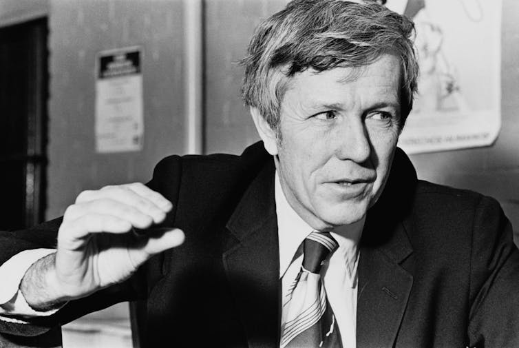 Black and white image of Harrington, seated in a suit and tie, speaking.