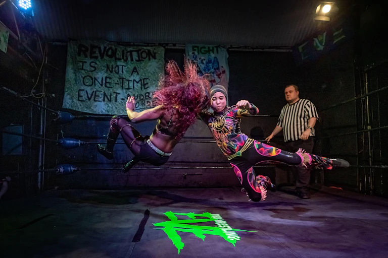 Two female wrestlers tackle each other during a match while referee stands in the background