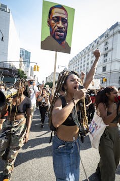 A woman with a fist in the air shouts into a microphone as she marches in a crowd in urban streets, a green and brown illustration of a man held aloft on a poster behind the woman.