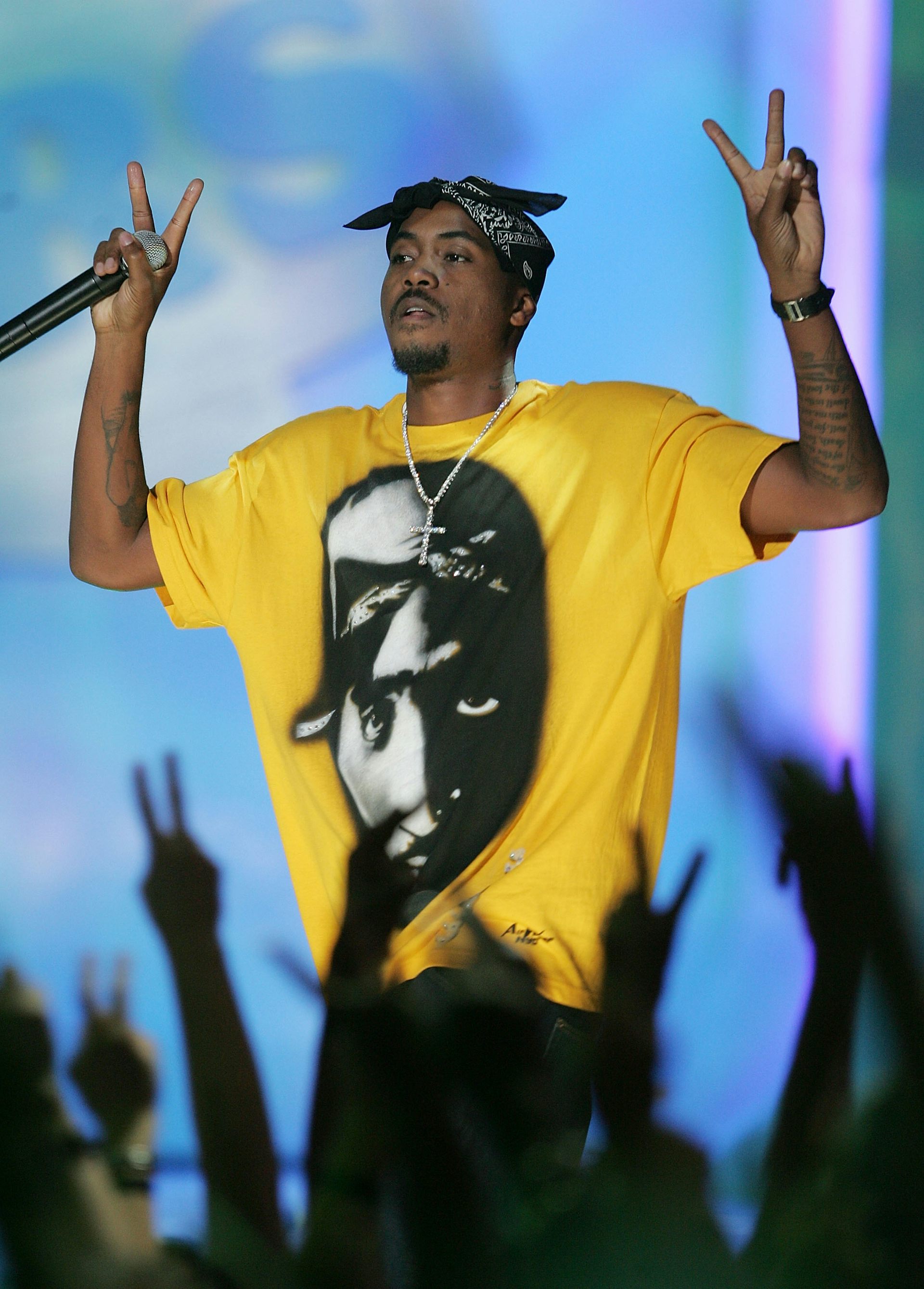 A man on stage raises both hands in peace signs as a crowd of arms from spectators do the same. He wears a yellow T-shirt with a prominent image of a man on it.