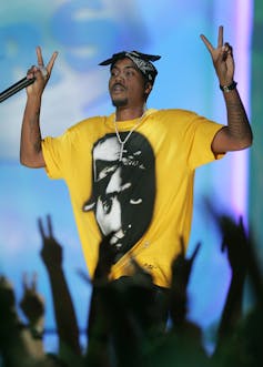 A man on stage raises both hands in peace signs as a crowd of arms from spectatotrs do the same. He wears a yellow T-shirt with a prominent image of a man on it.