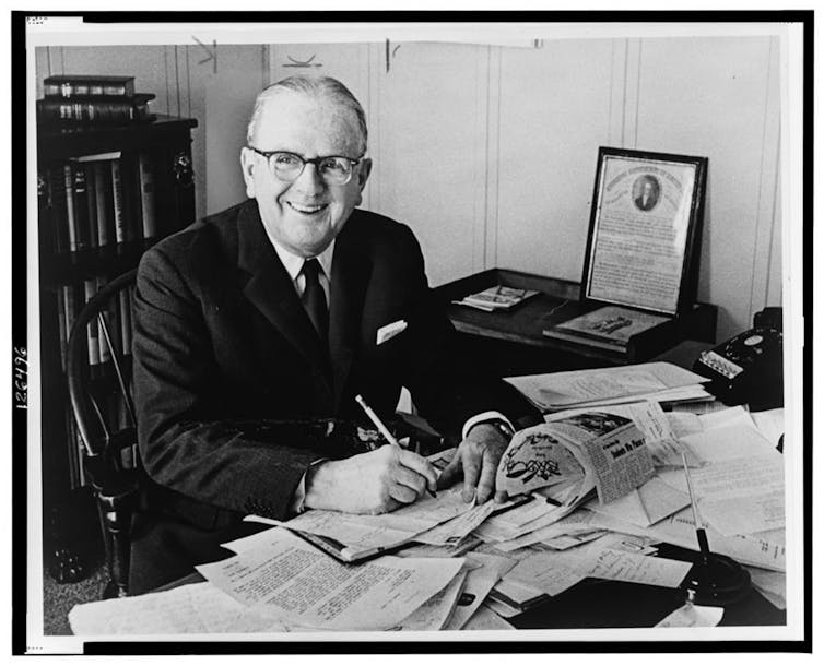 A man sits at a desk covered in paper.