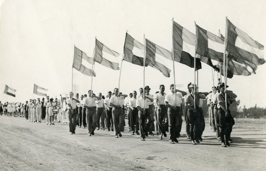 Men wearing shirts with rolled-up sleeves and long pants march with flags hoisted high 