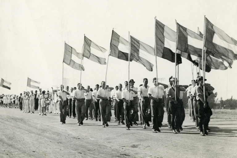 Men wearing shirts with rolled-up sleeves and long pants march with flags hoisted high