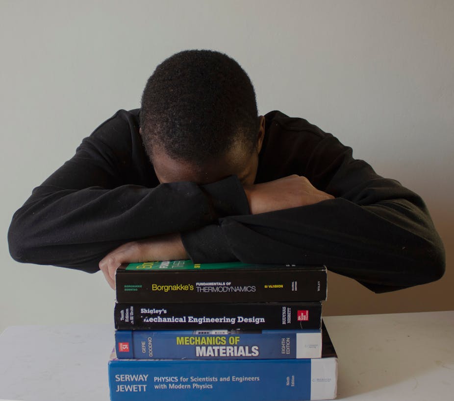 A man in a black shirt hides his face in his folded arms, poised atop a pile of what appear to be university textbooks.