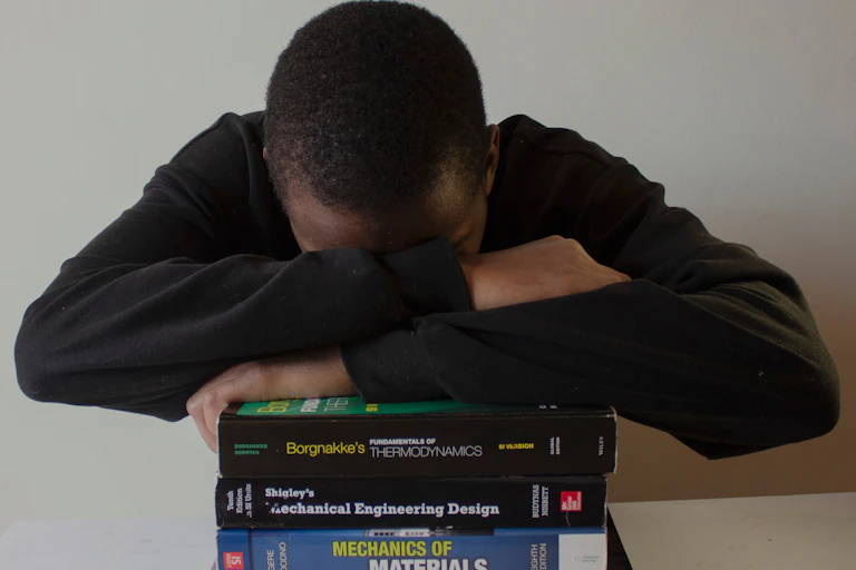 A man in a black shirt hides his face in his folded arms, poised atop a pile of what appear to be university textbooks.