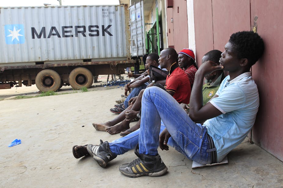 A group of young men seated outside a warehouse with a loading truck nearby.