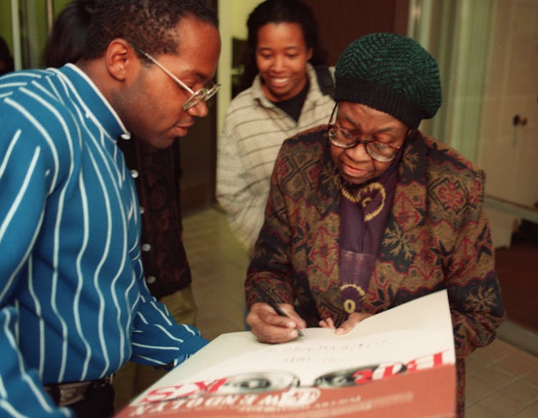 A Black woman wearng glasses and a green hat signs a poster.