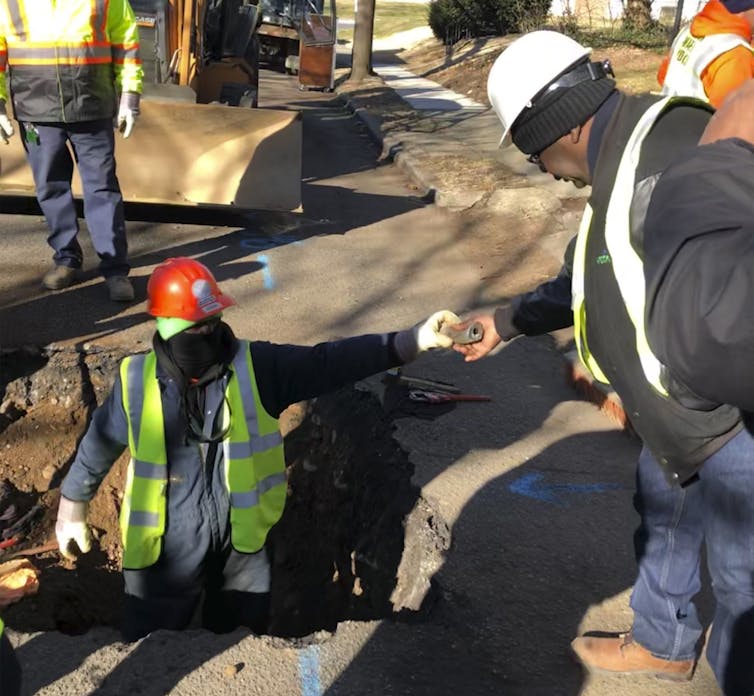 Worker standing in a trench dug in the street hands a piece of pipe to a colleague.