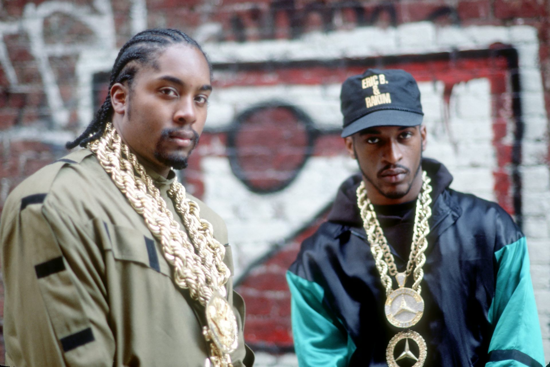 Two rappers wearing golden chains pose for a photo in front of a wall cover...