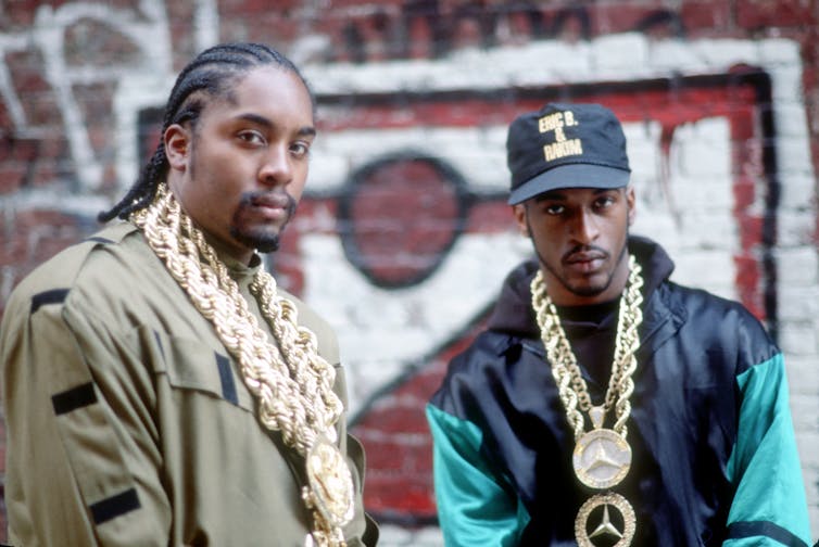 Dois rappers usando correntes douradas posam para uma foto em frente a uma parede coberta de pichações.