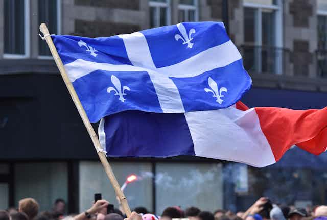 Un drapeau québécois et un drapeau français flottent au vent devant des édifices lors d'une manifestation
