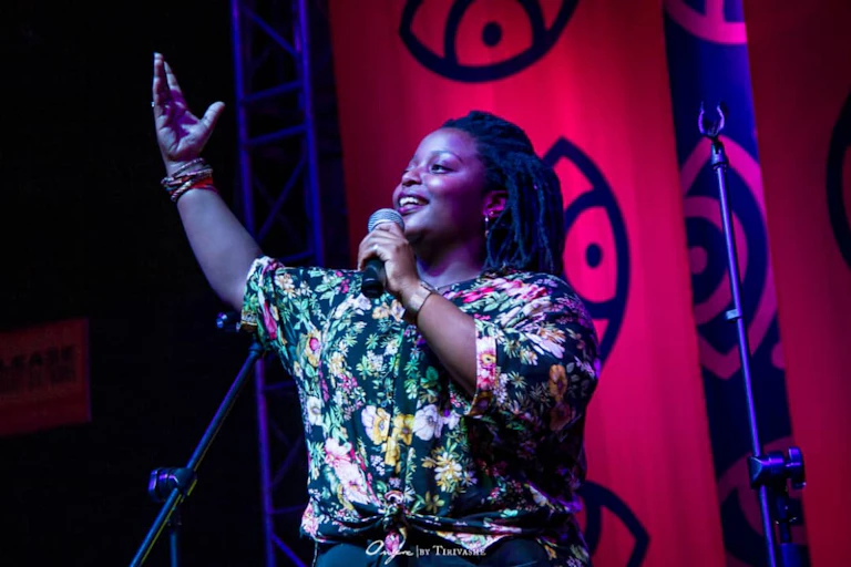 A woman stands on stage, a mic in her hand and her other arm raised, smiling as the light catches her face.