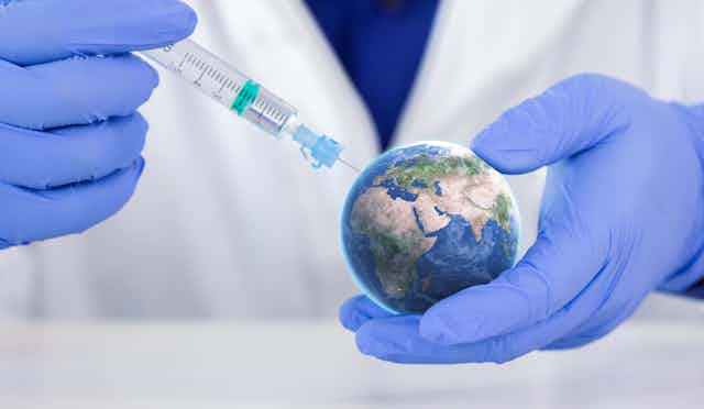 Un sanitario con guantes azules y bata blanca aplicando una inyección a una pequeña bola del mundo.