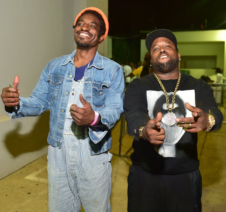 Two rappers pose for a photo in a hallway.