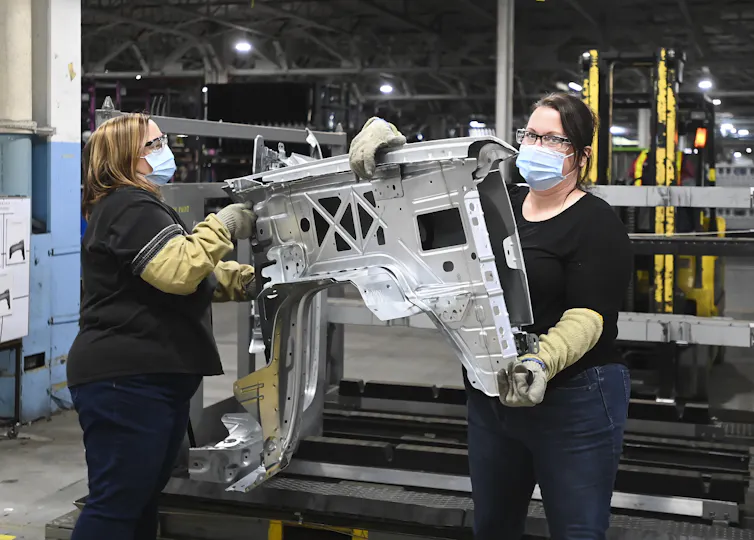 Two women GM workers carry truck fenders.