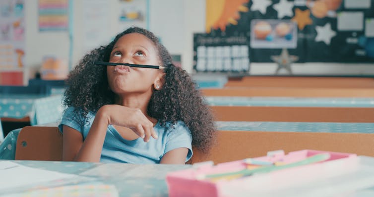 A young girl balances a pencil on her upper lip in class.