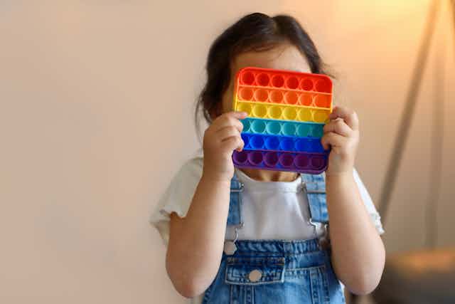 Little girl in white shirt and blue overalls holding a multicolored square popping toy in front of her face