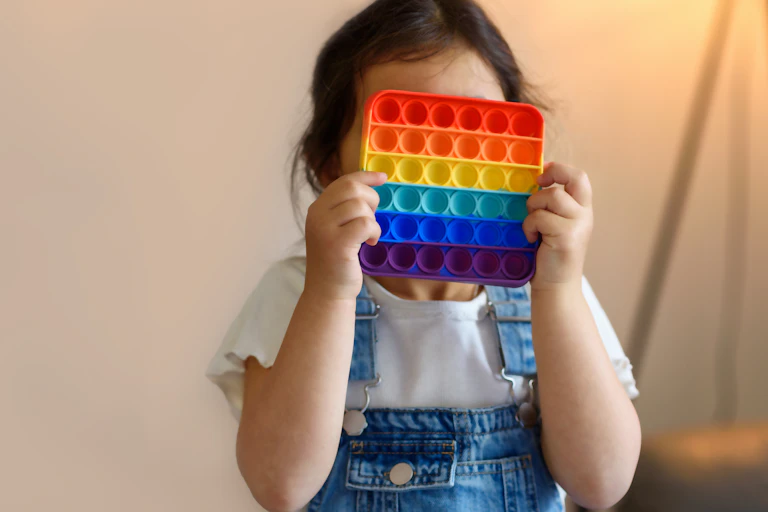 Little girl in white shirt and blue overalls holding a multicolored square popping toy in front of her face