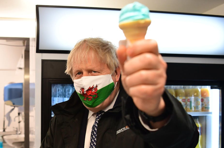 Boris Johnson holds up a blue ice cream while wearing a Welsh flag face mask. 