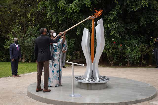 A man and a woman, both wearing face masks, hold a pole to light a flame on a monument in a garden. 