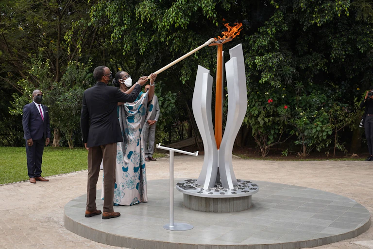 A man and a woman, both wearing face masks, hold a pole to light a flame on a monument in a garden.