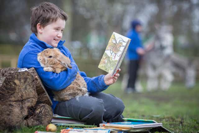 A boy in school uniform holds a big rabbit against his chest while reading in the playground
