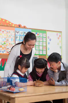 A teacher looks down at three children petting a guinea pig in the classroom