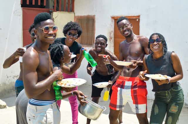 Seven young men and women dance in a backyard holding plates of food, smiling and having fun.