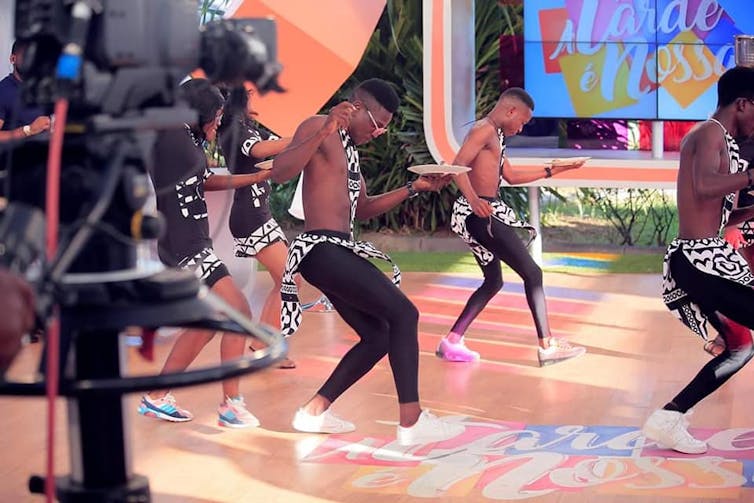 A television studio, a large camera foregrounded. In front of the cameras, a group of young men dances.