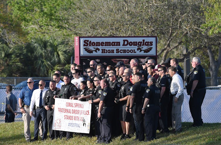 Group of uniformed police stand with a sign welcoming back students in front of the school, on a sunny day