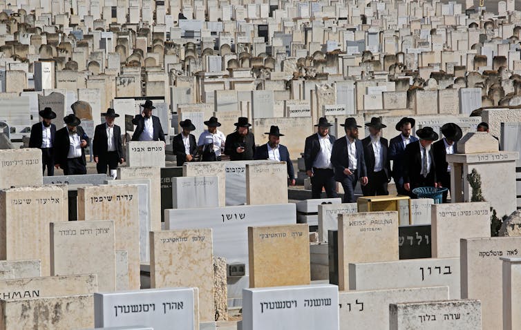 Bearded men dressed in black and wearing wide-brimmed black hats walk amid a sea of gravestones.