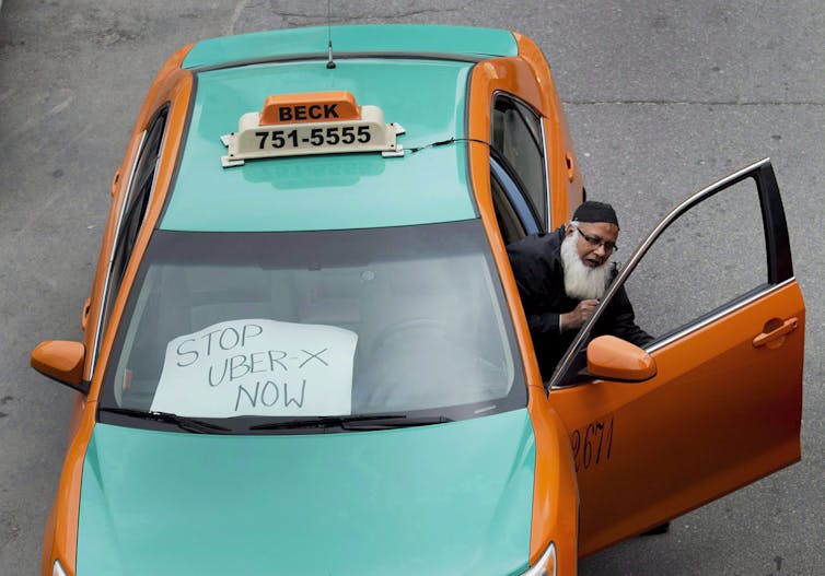 A taxi driver gets out of his car, which has a Stop Uber sign in its window.